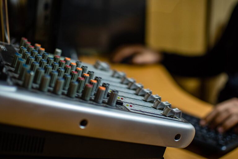 Un gros plan d'une console de mixage audio avec divers boutons et curseurs. En arrière-plan, une personne utilise un clavier.