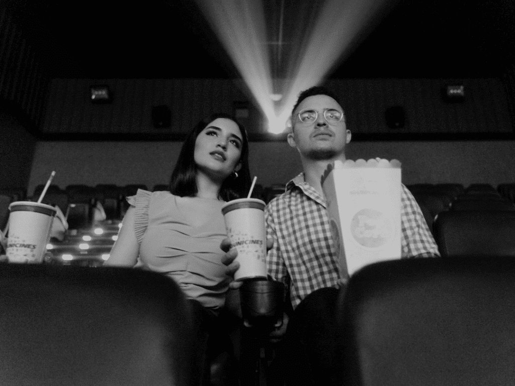 Un homme et une femme sont assis dans une salle de cinéma, tenant des boissons et du pop-corn, avec les faisceaux de lumière du projecteur visibles en arrière-plan.
