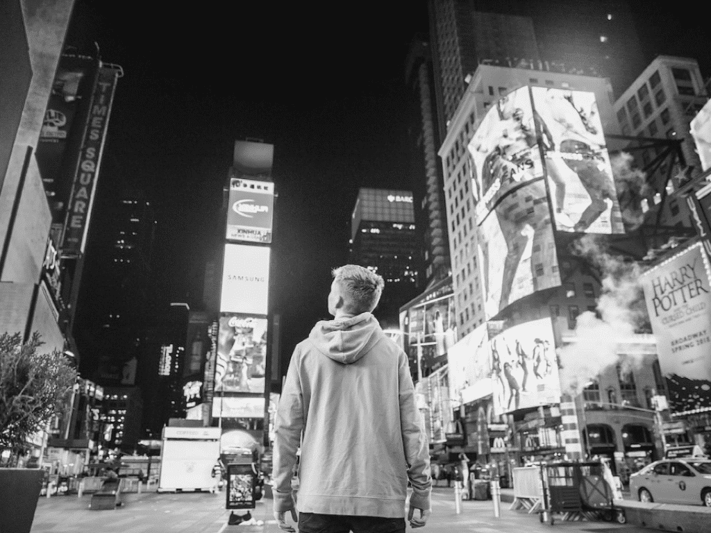 A person wearing a hoodie stands in the middle of a city street at night, looking up at brightly lit billboards and skyscrapers.