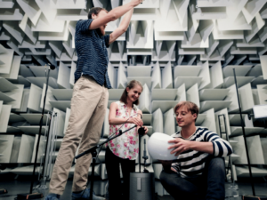 Three people are in an anechoic chamber. One is adjusting equipment overhead, one is holding a microphone, and one is kneeling, holding a white spherical object. The walls are covered in sound-absorbing panels.