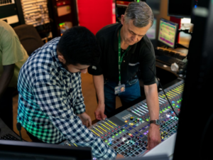 Deux hommes travaillant sur une grande console de mixage audio dans une salle de contrôle, ajustant divers boutons et curseurs, avec des écrans d'ordinateur en arrière-plan.