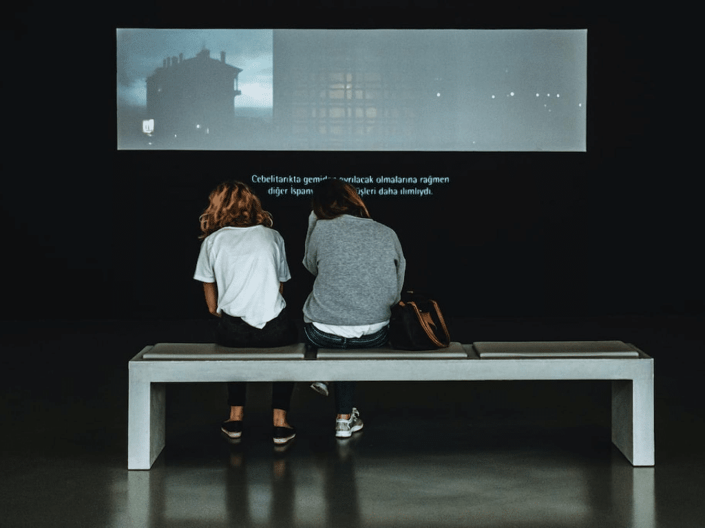 Deux personnes sont assises sur un banc dans une pièce sombre face à un grand écran affichant du texte et des images.