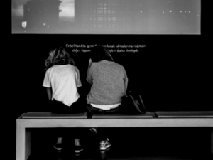 Deux personnes sont assises sur un banc face à un grand écran affichant une image avec du texte. Le décor semble être un espace d’exposition intérieur. La scène est capturée en noir et blanc.