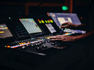 Mains ajustant les commandes sur une console de mixage audio professionnelle avec des écrans et des boutons bien éclairés dans un environnement faiblement éclairé.