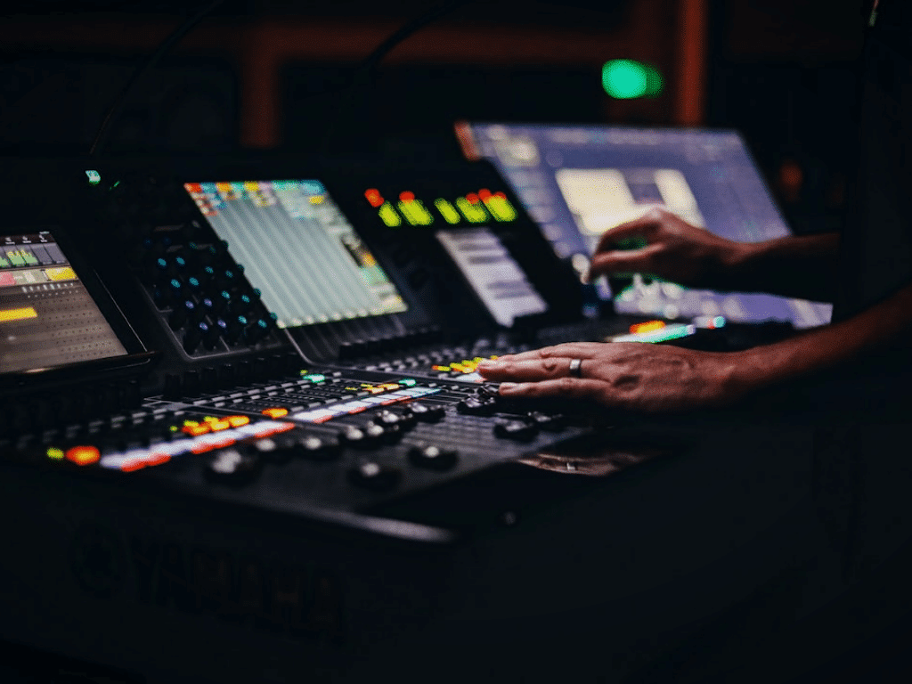Mains ajustant les commandes sur une console de mixage audio professionnelle avec des écrans et des boutons bien éclairés dans un environnement faiblement éclairé.