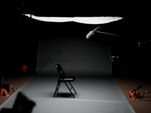 A dimly lit, professional photography or film studio setup with a single folding chair in the center, surrounded by lights and microphones.