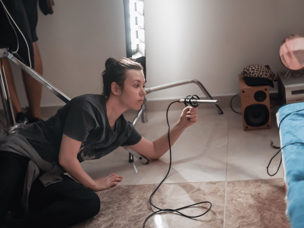 A person is positioned on the floor in a studio setting, holding a microphone and a cable. Nearby are a bright light, a blue object, and an orange speaker.