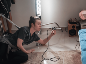 Une personne accroupie sur le sol tient un microphone pointé vers une couverture bleue, entourée d'équipements audio et d'éclairage.