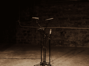 A microphone stand with two microphones, set against a dimly lit brick wall background on a stage.