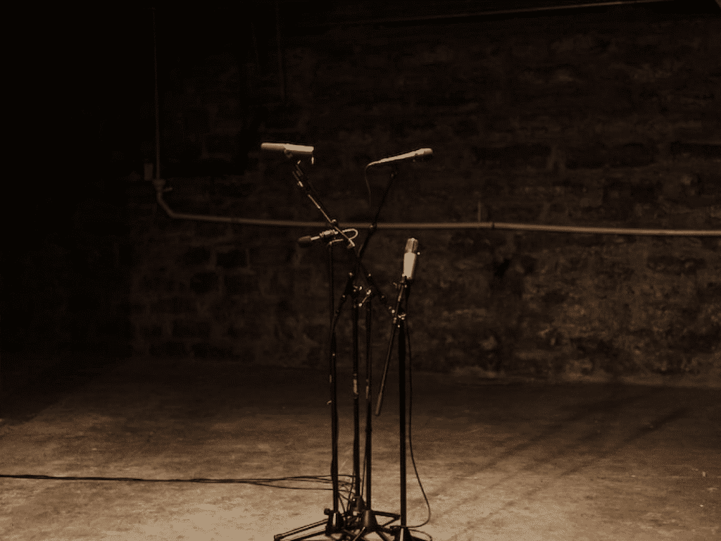 A microphone stand with two microphones, set against a dimly lit brick wall background on a stage.