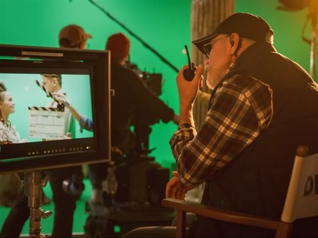 Director seated in front of a monitor with a clapboard displayed. Crew members in the background set up equipment against a green screen.
