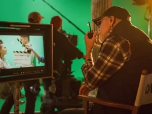 Un réalisateur portant une casquette et une chemise à carreaux observe un plateau de tournage derrière un moniteur, tenant un talkie-walkie, avec un écran vert en arrière-plan.