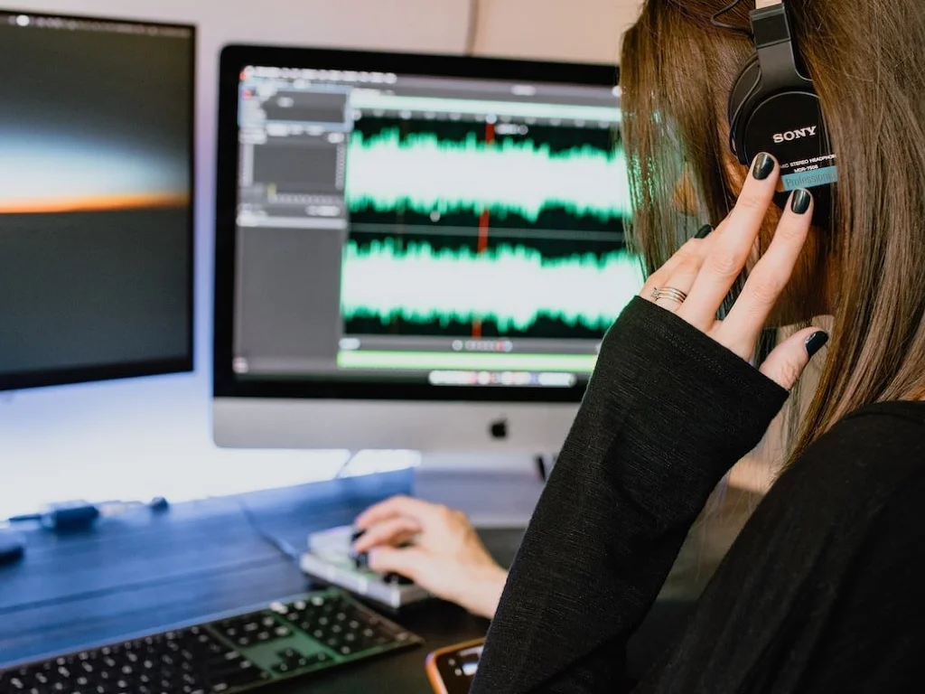 Person wearing Sony headphones working on audio editing software on a computer with two monitors displaying waveforms.