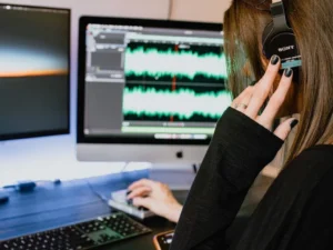 In a bustling dubbing studio, a person wearing headphones intently works at a computer, the screen alive with intricate audio editing software.