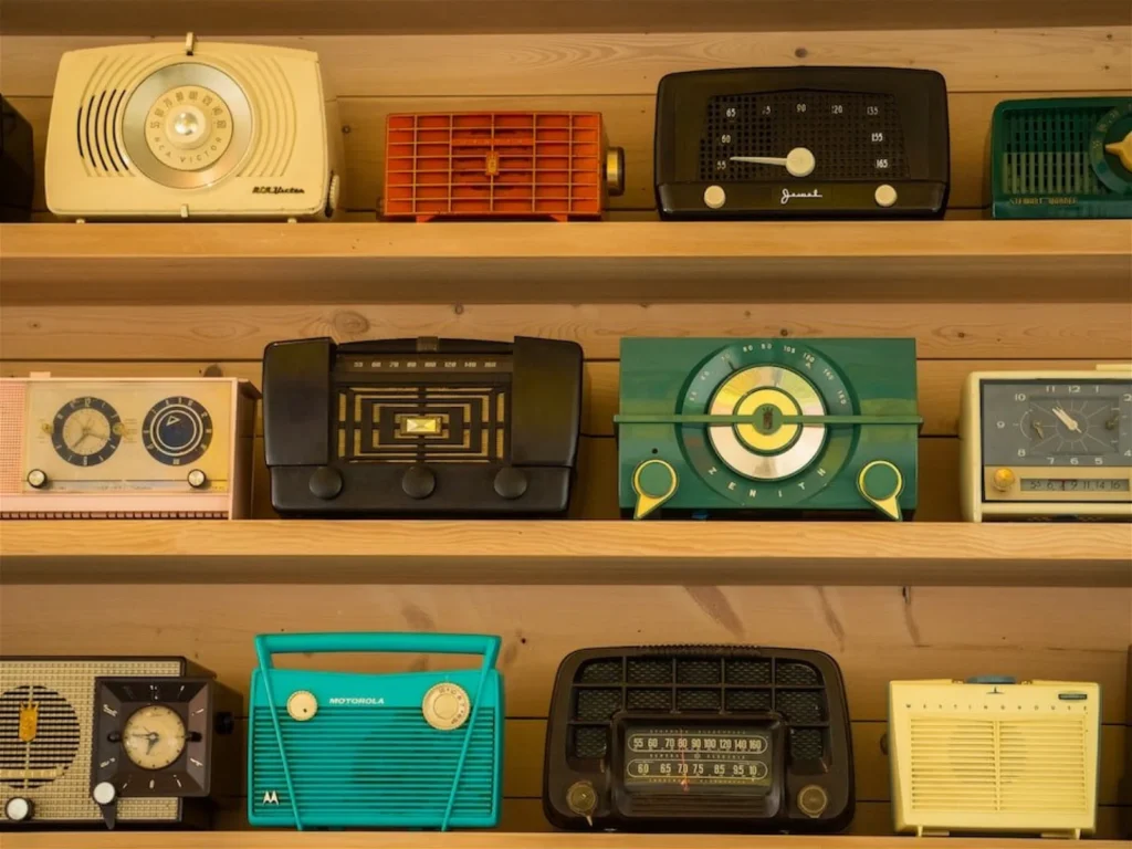 A collection of vintage radios is displayed on wooden shelves, featuring various designs and colors.