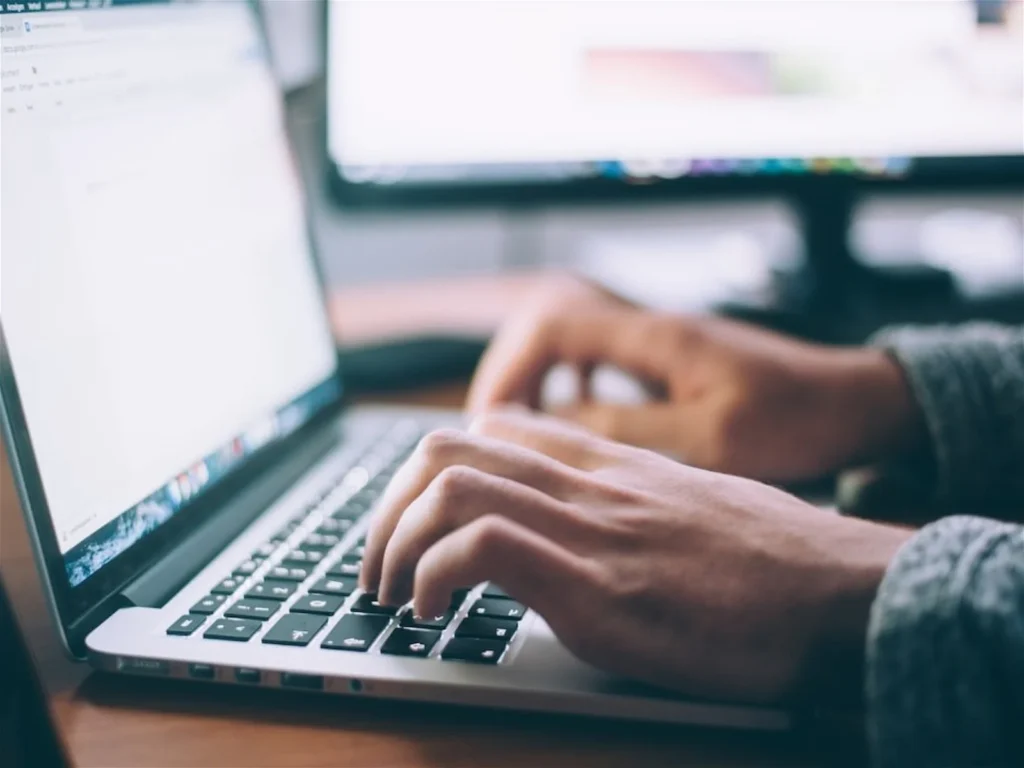 Person typing on a laptop keyboard with a blurred computer screen in the background.