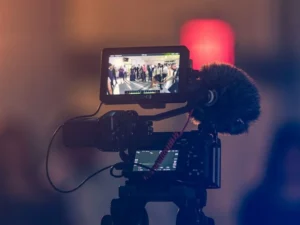 Video camera recording a group of people gathered indoors, displayed on its screen, with a blurred background and a warm light glow.