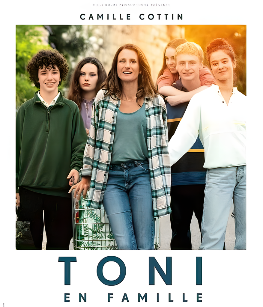 A woman with brown hair in a checkered shirt stands smiling in front of a group of five teenagers, some with arms around each other. The background shows greenery, reminiscent of a scene from a dubbing studio. Text: "Toni en Famille.