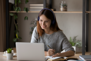 A woman wearing headphones smiles while working in her home dubbing studio. She uses a laptop at a desk, holding a pen with an open book in front of her. Shelves filled with plants and books create a cozy backdrop.