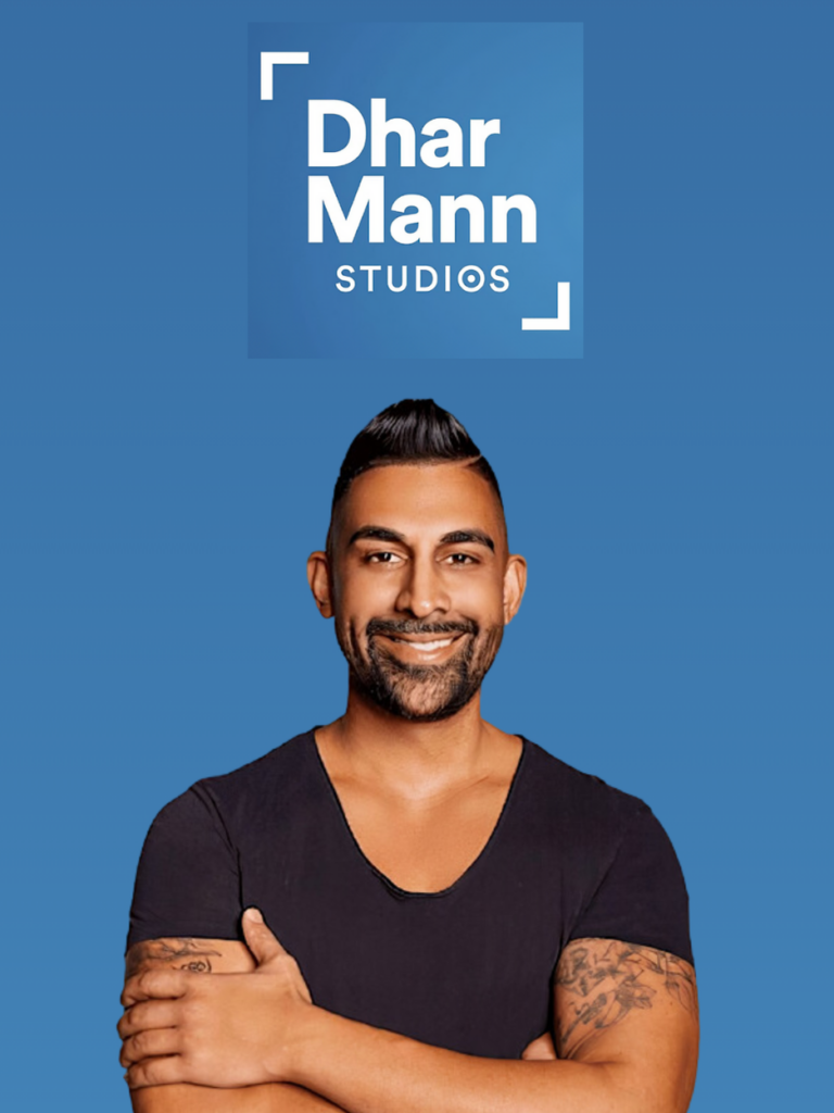 Homme barbu et tatoué, souriant et croisant les bras, avec un logo "dhar mann studios" au-dessus sur fond bleu pour un projet de collaboration.