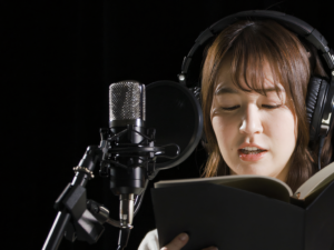 In a dubbing studio, a person wearing headphones reads from a book into a microphone, set against a dark background.