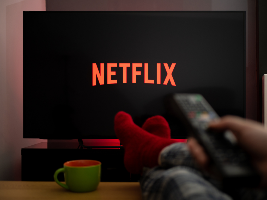 A person with red socks holds a remote, feet up, watching Netflix on a TV screen. A green mug sits on a table nearby.