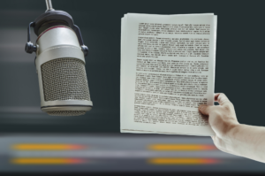 A person holds printed script pages near a large studio microphone in a bustling dubbing studio, set against a blurred background with an orange-striped table.