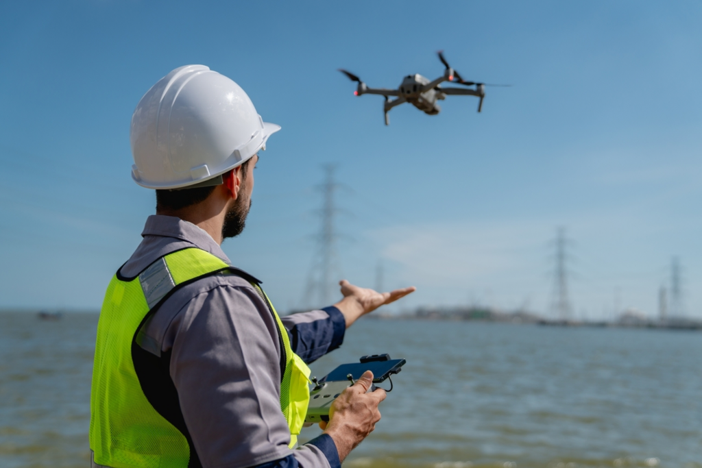 A person in a hard hat and high-visibility vest operates a drone near water, holding a controller with precision akin to an engineer in a dubbing studio. Power lines and pylons stretch across the background under a clear blue sky.