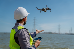 A person in a hard hat and high-visibility vest operates a drone near water, holding a controller with precision akin to an engineer in a dubbing studio. Power lines and pylons stretch across the background under a clear blue sky.