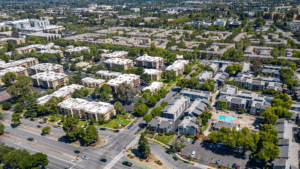 Aerial drone view of a suburban neighborhood with multiple apartment complexes, tree-lined streets, parking lots, a main road, and a swimming pool visible among the buildings.