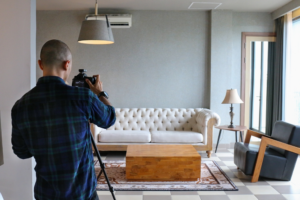 A person photographing an empty living room with a dubbing studio vibe, featuring a white sofa, wooden coffee table, and black armchair.