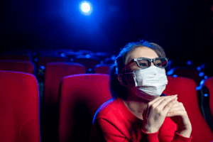 A person wearing 3D glasses and a face mask sits in a red theater seat, watching séries chinoises in a dimly lit, mostly empty cinema, reflecting adaptation culturelle.
