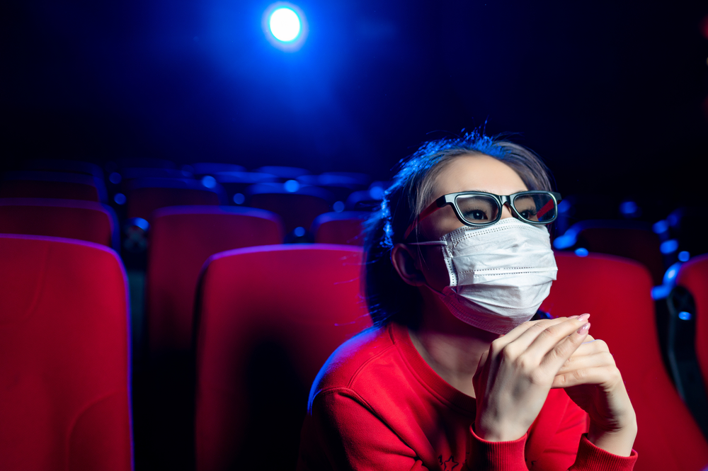 A person wearing 3D glasses and a face mask sits in a red theater seat, watching séries chinoises in a dimly lit, mostly empty cinema, reflecting adaptation culturelle.
